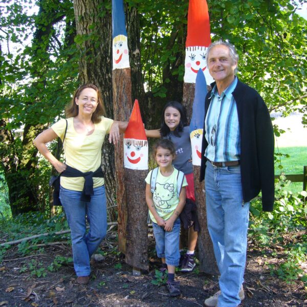 Vier Personen stehen im Freien in der Nähe von Bäumen und drei hohen Holzstatuen, die als lächelnde Gnome mit roten und blauen Hüten bemalt sind.