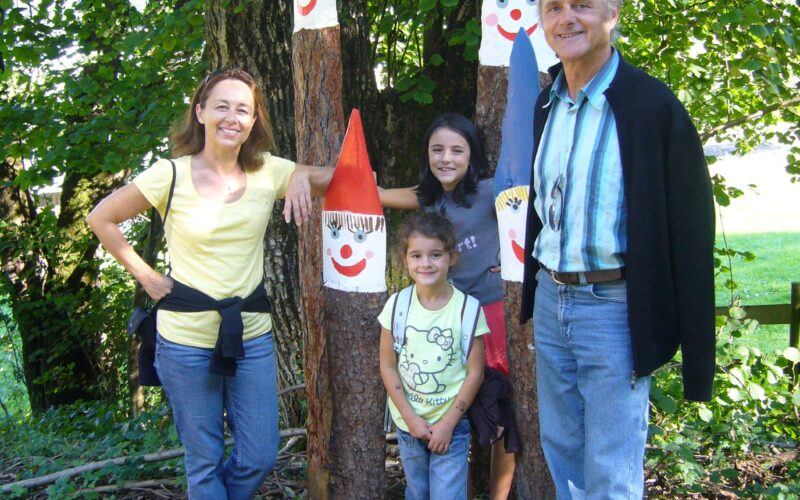 Vier Personen stehen im Freien in der Nähe von Bäumen und drei hohen Holzstatuen, die als lächelnde Gnome mit roten und blauen Hüten bemalt sind.