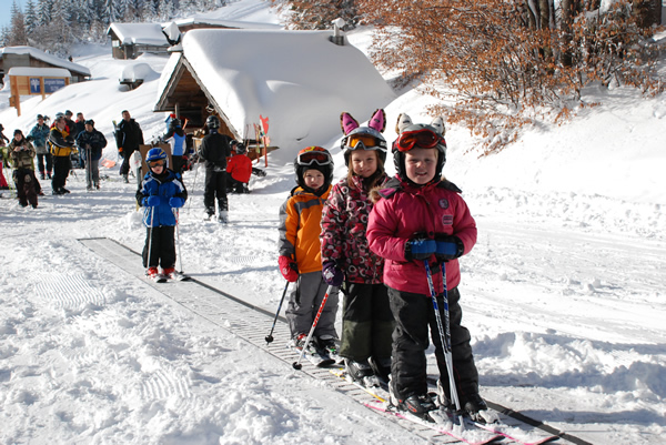 Kinder in Winterkleidung stehen in einer Reihe auf einem Skiförderband, im Hintergrund sind schneebedeckte Hütten und andere Menschen zu sehen.