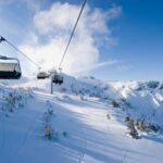 Ein Skilift befördert leere Sessel über eine verschneite Berglandschaft mit Bäumen und einem strahlend blauen Himmel.
