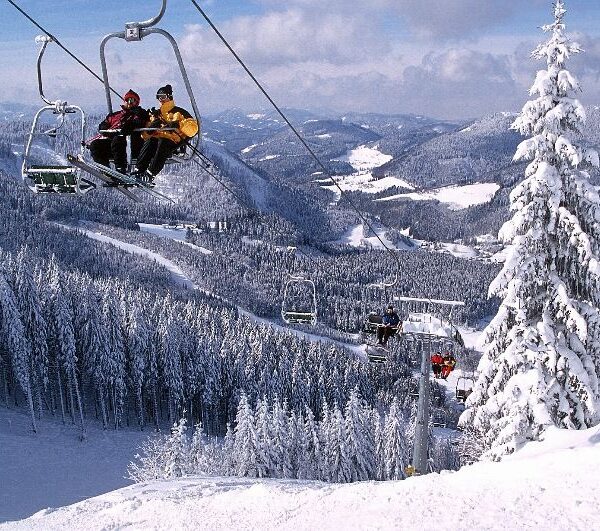 Menschen fahren mit einem Skilift über eine verschneite Berglandschaft mit schneebedeckten Bäumen und Hügeln im Hintergrund.