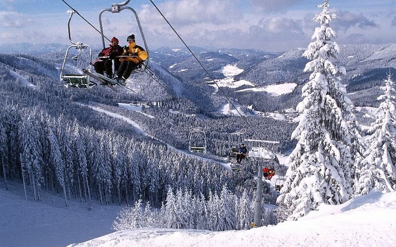 Menschen fahren mit einem Skilift über eine verschneite Berglandschaft mit schneebedeckten Bäumen und Hügeln im Hintergrund.