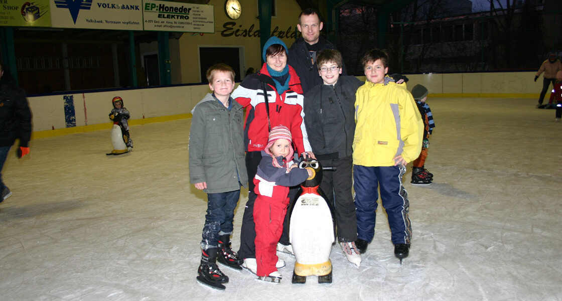 Sechs Kinder und ein Erwachsener posieren gemeinsam auf einer Eislaufbahn; ein Kind hält einen Pinguin als Eislaufhilfe.