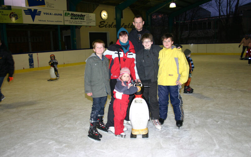 Sechs Kinder und ein Erwachsener posieren gemeinsam auf einer Eislaufbahn; ein Kind hält einen Pinguin als Eislaufhilfe.