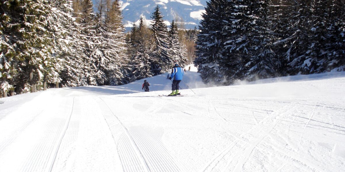 Zwei Personen fahren auf einer präparierten, schneebedeckten Piste, umgeben von schneebedeckten immergrünen Bäumen, unter einem klaren Himmel.