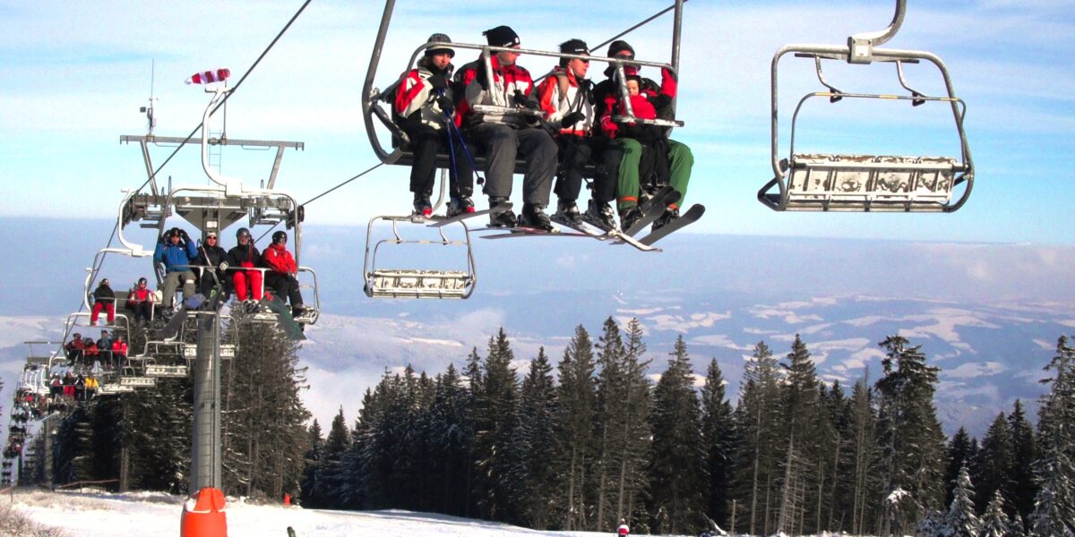 Skifahrer in roten Jacken fahren mit einem Skilift über eine verschneite Piste, unter denen sich Skifahrer und Snowboarder befinden, die von Bäumen und Bergen umgeben sind.