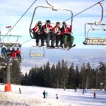 Skifahrer in roten Jacken fahren mit einem Skilift über eine verschneite Piste, unter denen sich Skifahrer und Snowboarder befinden, die von Bäumen und Bergen umgeben sind.