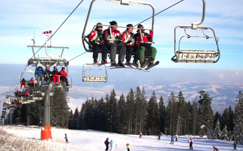 Skifahrer in roten Jacken fahren mit einem Skilift über eine verschneite Piste, unter denen sich Skifahrer und Snowboarder befinden, die von Bäumen und Bergen umgeben sind.