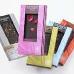 Fünf verschiedene Bachhalm-Schokoladentafeln in einer bunten Verpackung, übereinander angeordnet, mit Fenstern, die die verschiedenen Schokoladensorten im Inneren zeigen.