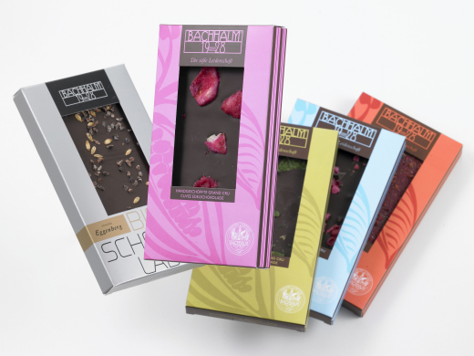Fünf verschiedene Bachhalm-Schokoladentafeln in einer bunten Verpackung, übereinander angeordnet, mit Fenstern, die die verschiedenen Schokoladensorten im Inneren zeigen.