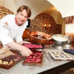 Ein Koch arrangiert Tabletts mit bunten Pralinen auf einer Theke, während in einer dekorierten Küche Schalen mit Zutaten und Formen zu sehen sind.