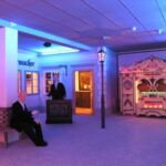 Eine schwach beleuchtete Innenraumszene mit Schaufensterpuppen, einer alten Orgel mit Lichtern, einer Bank und altmodischen Schaufenstern bei blau-violetter Beleuchtung.