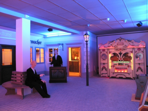 Eine schwach beleuchtete Innenraumszene mit Schaufensterpuppen, einer alten Orgel mit Lichtern, einer Bank und altmodischen Schaufenstern bei blau-violetter Beleuchtung.