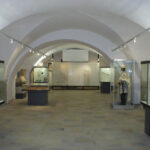 Eine Museumsgalerie mit gewölbten Decken, Vitrinen mit Artefakten und einer Schaufensterpuppe in historischer Kleidung auf der rechten Seite.