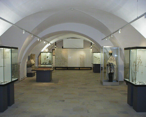 Eine Museumsgalerie mit gewölbten Decken, Vitrinen mit Artefakten und einer Schaufensterpuppe in historischer Kleidung auf der rechten Seite.