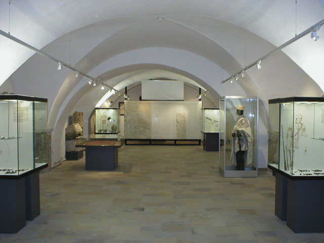 Eine Museumsgalerie mit gewölbten Decken, Vitrinen mit Artefakten und einer Schaufensterpuppe in historischer Kleidung auf der rechten Seite.