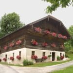 Ein zweistöckiges Haus mit hölzernen oberen Wänden und weißen unteren Wänden, verziert mit rosafarbenen Blumenkästen an den Fenstern, liegt zwischen zwei kurvenreichen Straßen, umgeben von Grün.