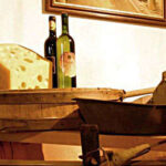 Ein großes Stück Käse, zwei Weinflaschen und rustikale Küchenutensilien sind auf einer hölzernen Fläche angeordnet, dahinter hängt ein gerahmtes Bild an der Wand.