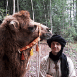 Ein Mann mit Kopftuch steht neben einem Kamel in einem Wald mit Bäumen und Grünpflanzen im Hintergrund.