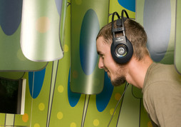 Ein Mann mit Kopfhörern lehnt sich an einen kleinen Bildschirm, der an einer bunten, gemusterten Wand angebracht ist.