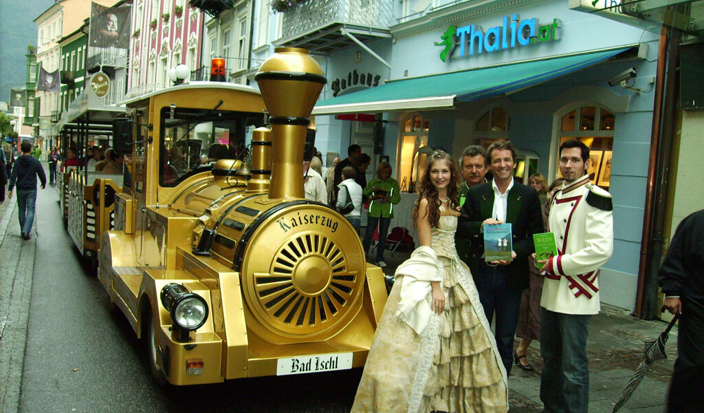 Eine Gruppe formell gekleideter Personen steht neben einem goldfarbenen Touristenzug in einer von Geschäften, darunter Thalia.at, gesäumten Straße in Bad Ischl, Österreich.