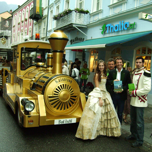 Eine Gruppe formell gekleideter Personen steht neben einem goldfarbenen Touristenzug in einer von Geschäften, darunter Thalia.at, gesäumten Straße in Bad Ischl, Österreich.