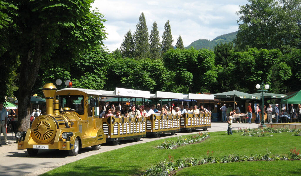 Ein kleiner gelber Touristenzug voller Fahrgäste fährt durch einen Park mit grünen Rasenflächen, Blumenbeeten und Bäumen. Im Hintergrund sind Marktstände und Menschen zu sehen.