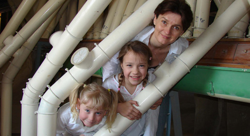 Ein Erwachsener und zwei junge Mädchen lächeln und posieren zwischen weißen Kunststoffrohren in einer industriellen oder mechanischen Umgebung.