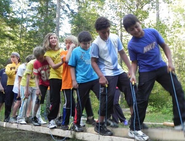 Eine Gruppe von Kindern steht eng beieinander auf einer Holzplanke im Freien, hält sich an Seilen fest und balanciert im Rahmen einer Gruppenaktivität in einem Waldgebiet.
