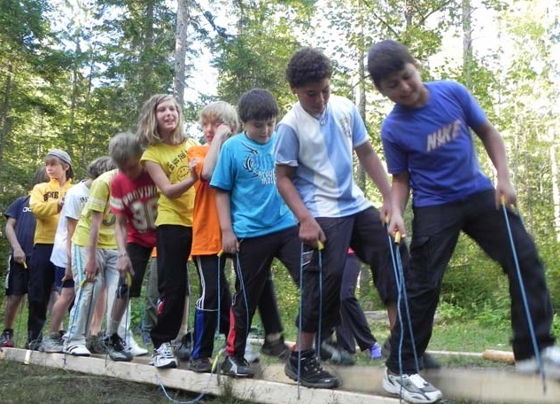 Eine Gruppe von Kindern steht eng beieinander auf einer Holzplanke im Freien, hält sich an Seilen fest und balanciert im Rahmen einer Gruppenaktivität in einem Waldgebiet.
