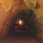 Ein schwach beleuchteter unterirdischer Tunnel mit gewölbten Decken und zerkratzten Steinwänden, der zu einem von einer einzigen Lampe beleuchteten Eingangstor führt.