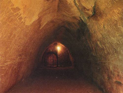 Ein schwach beleuchteter unterirdischer Tunnel mit gewölbten Decken und zerkratzten Steinwänden, der zu einem von einer einzigen Lampe beleuchteten Eingangstor führt.