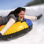 Eine Frau in Winterkleidung rutscht lächelnd auf einem bunten, aufblasbaren Schlauch einen verschneiten Hügel hinunter, während im Hintergrund andere Leute Schlauchboot fahren.