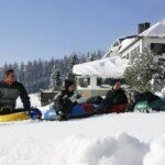 Vier Personen sitzen auf Snow Tubes in einer verschneiten Landschaft in der Nähe eines großen Gebäudes, mit schneebedeckten Bäumen und einem klaren Himmel im Hintergrund.