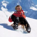 Eine Person in einer rot-schwarzen Jacke schlittelt bergab auf Schnee mit Bergen im Hintergrund.