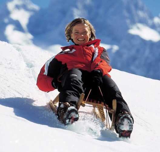 Eine Person in einer rot-schwarzen Jacke schlittelt bergab auf Schnee mit Bergen im Hintergrund.