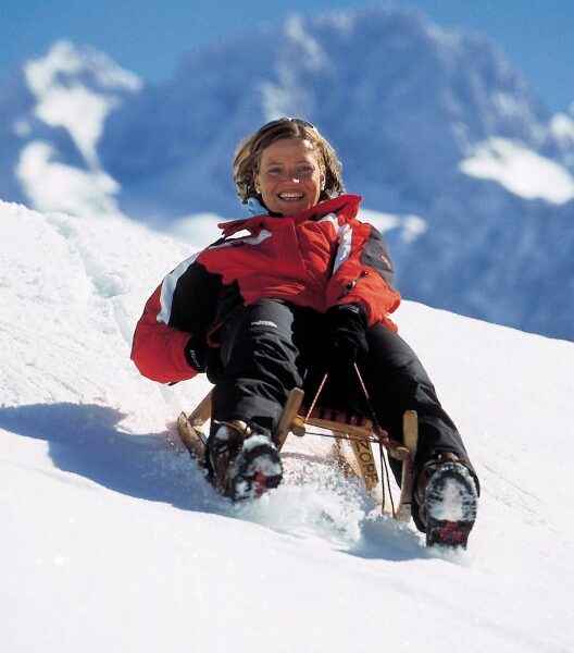 Eine Person in einer rot-schwarzen Jacke schlittelt bergab auf Schnee mit Bergen im Hintergrund.