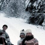 Drei Personen in Winterkleidung sitzen auf einem Schlitten in einem verschneiten Wald und lächeln in die Kamera. Schnee bedeckt die Bäume und den Boden um sie herum.
