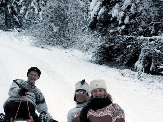 Drei Personen in Winterkleidung sitzen auf einem Schlitten in einem verschneiten Wald und lächeln in die Kamera. Schnee bedeckt die Bäume und den Boden um sie herum.