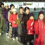 Eine Gruppe von Kindern und zwei Erwachsene stehen in einer Schlange auf einer Eislaufbahn, alle tragen Winterkleidung und Schlittschuhe.