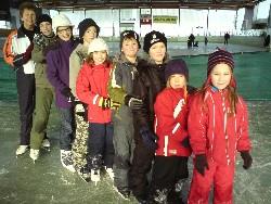 Eine Gruppe von Kindern und zwei Erwachsene stehen in einer Schlange auf einer Eislaufbahn, alle tragen Winterkleidung und Schlittschuhe.