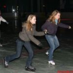 Drei junge Leute, die in einer Halle Schlittschuh laufen. Zwei Mädchen halten sich an den Händen und bewegen sich vorwärts, während ein anderes im Hintergrund Schlittschuh läuft.