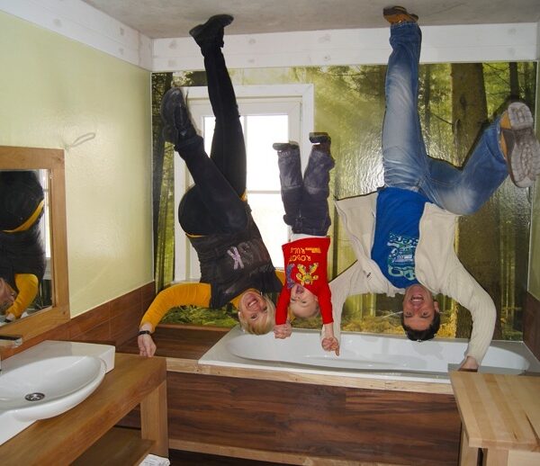 Drei Personen erscheinen kopfüber in einem Badezimmer mit einem Waldwandgemälde; der Raum ist so gestaltet, dass die Decke dem Boden ähnelt.