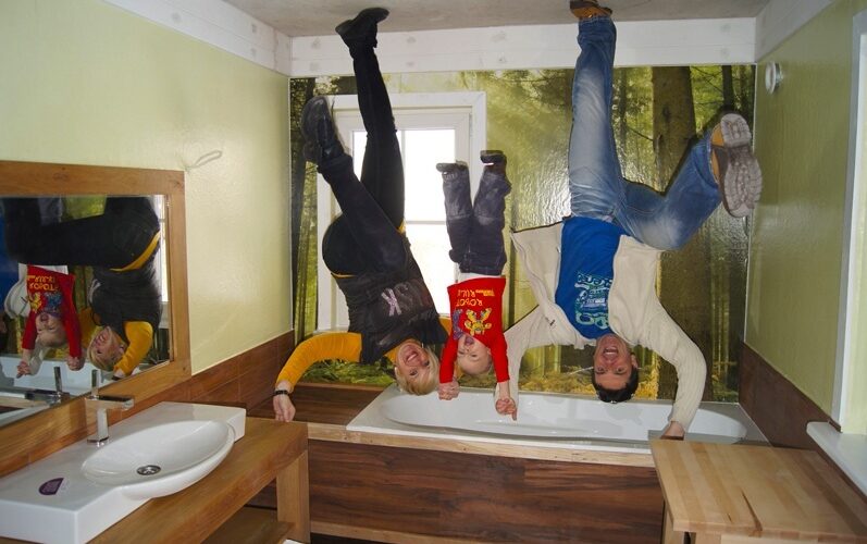 Drei Personen erscheinen kopfüber in einem Badezimmer mit einem Waldwandgemälde; der Raum ist so gestaltet, dass die Decke dem Boden ähnelt.