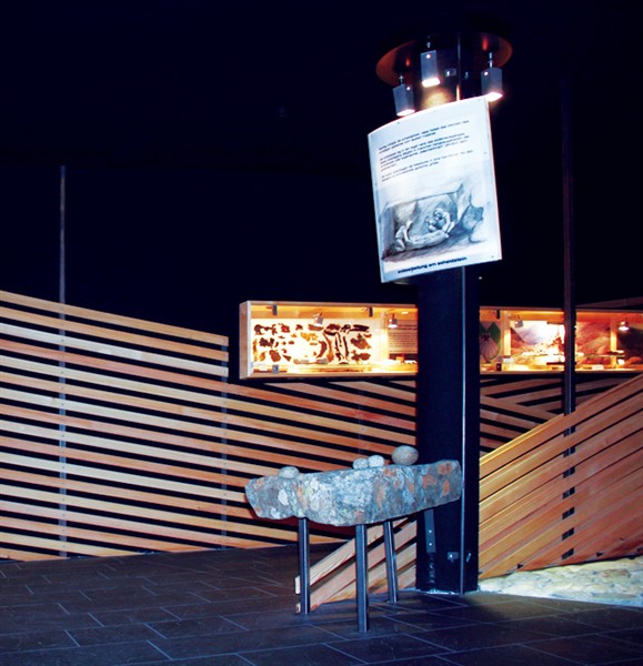 Eine Museumsausstellung zeigt ein Steinartefakt auf einem Ständer, eine Schautafel mit einem Bild und Text darüber und beleuchtete Vitrinen im Hintergrund.