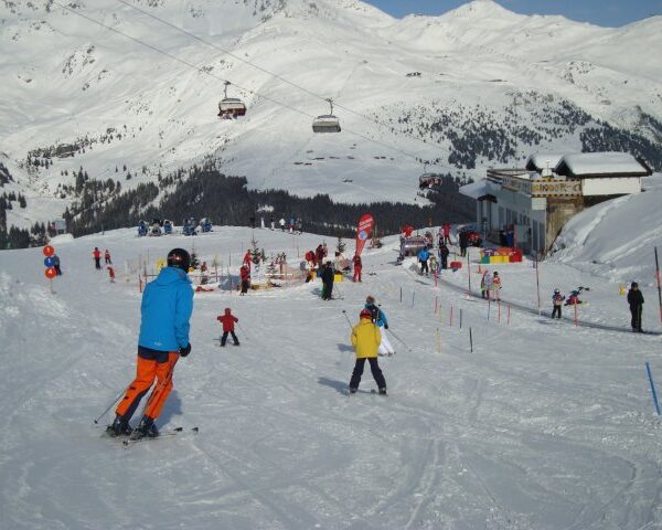 Skifahrer unterschiedlichen Alters fahren einen verschneiten Hang in der Nähe eines Skilifts hinunter, im Hintergrund sind eine Berglandschaft und ein Gebäude zu sehen.