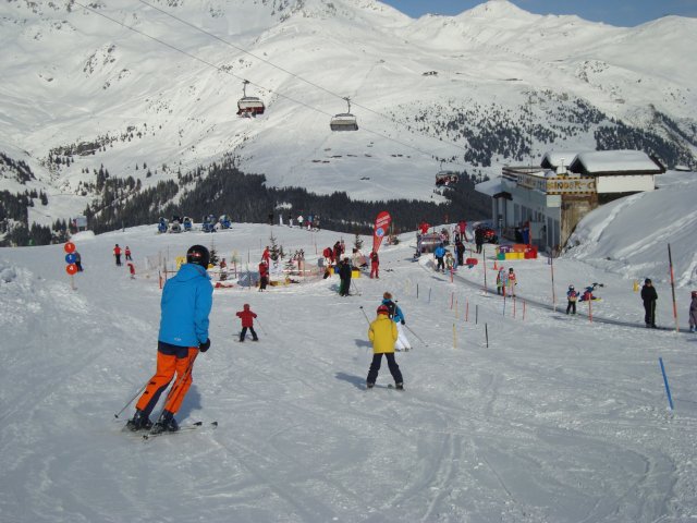 Skifahrer unterschiedlichen Alters fahren einen verschneiten Hang in der Nähe eines Skilifts hinunter, im Hintergrund sind eine Berglandschaft und ein Gebäude zu sehen.