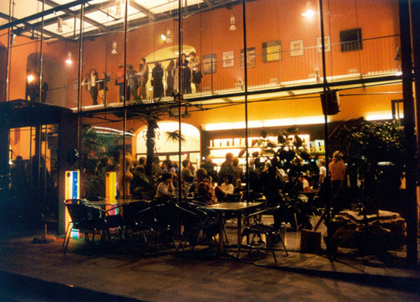 Ein modernes, gläsernes Café am Abend, in dem sich die Leute drinnen und auf dem oberen Balkon treffen, während andere an Tischen im Freien sitzen.