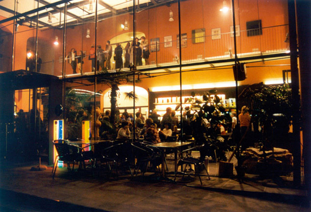 Ein modernes, gläsernes Café am Abend, in dem sich die Leute drinnen und auf dem oberen Balkon treffen, während andere an Tischen im Freien sitzen.