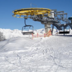 Eine Skiliftstation auf einem verschneiten Berg mit leeren Sesseln, schneebedecktem Boden und entfernten Bergen unter einem klaren blauen Himmel.